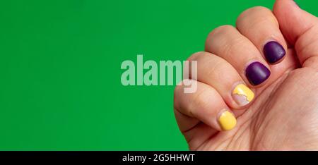 Die Nägel an der Hand mit beschädigtem Nagellack sind lila und gelb. Grüner Hintergrund mit Platz für Text. Selektiver Fokus. Stockfoto