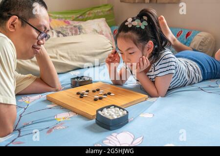 Asiatische niedliche Kind Mädchen spielen Go oder chinesische traditionelle Brettspiel auf dem Bett, das Mädchen denkt, und Vater wartet auf ihren Umzug. Stockfoto