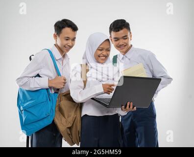 Drei Jugendliche in Schuluniformen lächeln mit einem Laptop zusammen, während sie einen Rucksack und ein Buch tragen Stockfoto