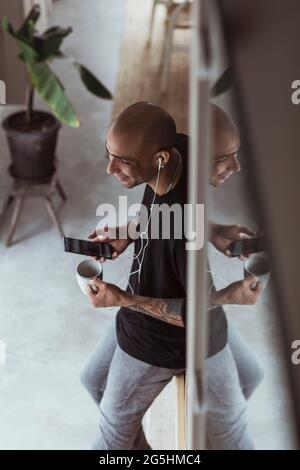 Blick aus der Ferne auf den Mann, der auf dem Mobiltelefon spricht, während er die Kaffeetasse zu Hause hält Stockfoto