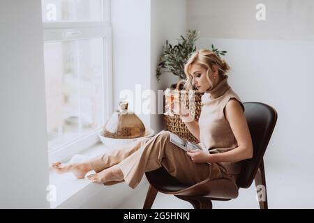 Junge Geschäftsfrau arbeitet zu Hause. Das nachdenkliche, ruhige Model hält ein Glas Weißwein und liest ein Magazin. Schöne Frau in einem modernen Interieur. Weiches Sel Stockfoto