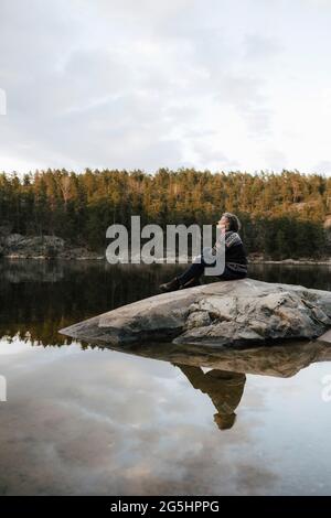 Weibliche Wanderin, die auf Felsen am See gegen den Himmel sitzt Stockfoto