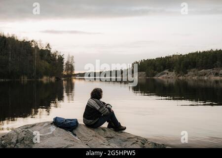 Weibliche Wanderer bewundern den See, während sie auf einem Felsen gegen den Himmel sitzt Stockfoto