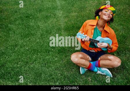 Rastafarian Mischrassen Frau sitzt auf grünem Gras und spielt blaue Ukulele. Junge Erwachsene mit Vitiligo genießen das Leben und die Freiheit Stockfoto