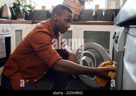 Junger afrikanischer Mann, der zuhause Kleidung in eine Waschmaschine legt Stockfoto