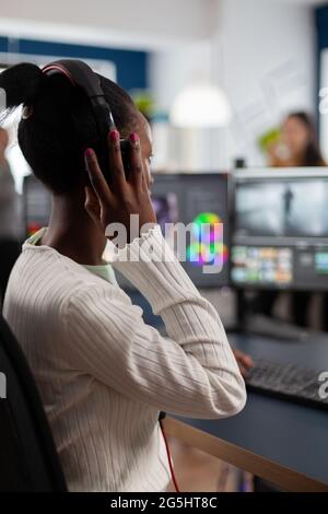 Schwarzer Videoeditor, der mit Filmmaterial auf dem pc mit zwei Monitoren arbeitet und Kopfhörer aufsetzt. Afrikanischer Videofilmer, der mit Post-Production-Software Filme bearbeitet, die in einem Büro der Kreativagentur arbeitet. Stockfoto