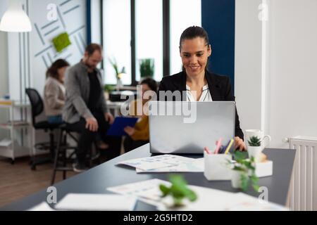 Glückliche hispanische Dame, die auf dem Laptop sitzt am Schreibtisch im Start-up-Büro Kaffee trinkt, während verschiedene Kollegen im Hintergrund arbeiten. Multiethnische Mitarbeiter planen ein neues Finanzprojekt. Stockfoto