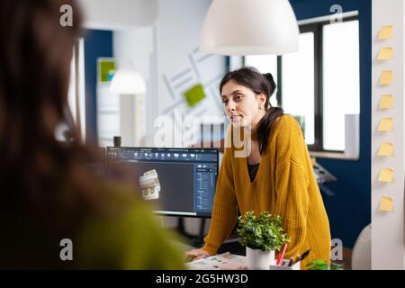Frau Industriedesignerin diskutiert mit Kollegin, die am Schreibtisch steht, während sie im CAD-Programm arbeitet und 3D-Prototypen von Komponenten entwirft. Ingenieur, der Ideen für Konstruktionsmechanismen teilt Stockfoto