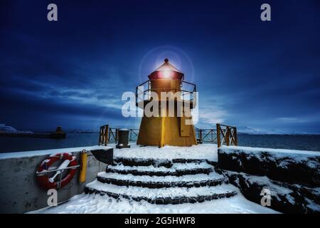 Leuchtturm am Eingang zum Alten Hafen, Reykjavik, Island. Im Winter schoss die blaue Stunde bei Sonnenaufgang, mit gelbem Leuchtfeuer und Schnee auf dem Boden. Stockfoto