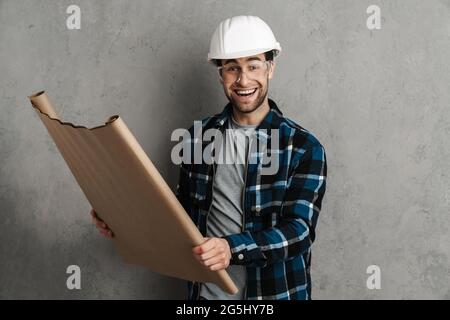Junger, glücklicher und hübscher Bauingenieur in Hardhut mit Blaupausen, die isoliert auf grauem Hintergrund stehen Stockfoto