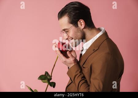 Glücklich mittleren Alters Brünette Mann in Jacke riechend rot Rose über rosa Wand Hintergrund Stockfoto