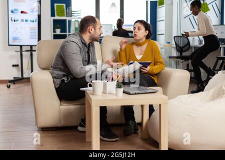 Verärgerter Mann Manager argumentiert Frau Angestellter für schlechte Arbeitsergebnisse, sitzen auf der Couch, während verschiedene Kollegen arbeiten Angst im Hintergrund. Geschäftsfrau Angestellter wütete über Multitasking schwierige Arbeit. Stockfoto