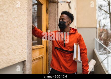 Junger Mann mit Schutzmaske, der mit Paketen vor dem Haus steht Stockfoto