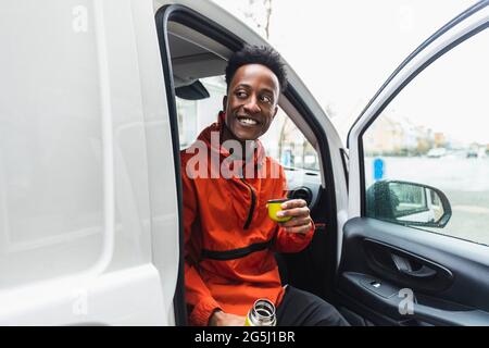 Lächelnder junger, unverzichtbarer Service-Mann, der mit einem Getränk im Lieferwagen dasaß und wegschaute Stockfoto