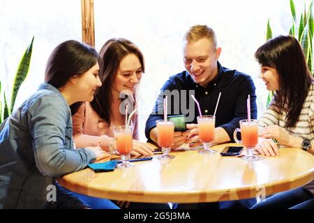 Ein Mann und ein Mädchen an einem Tisch in einem Café schauen auf ein Mobiltelefon - EINE Gruppe junger Menschen macht sich in einer Bar Spaß und nutzt ein Smartphone - die Conce Stockfoto