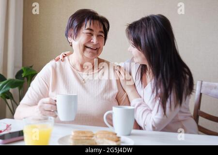 Glückliche junge Tochter umarmt eine ältere Mutter beim Frühstück am Tisch - zwei Generationen drücken Fürsorge und Wiedervereinigung aus - das Konzept o Stockfoto