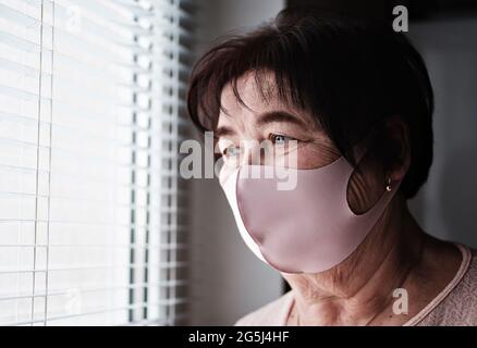 Eine ältere Frau, die eine Maske trägt, schaut während der Coronavirus-Epidemie traurigerweise aus dem Fenster - Oma bleibt während COVID-19 - Selbstisol zu Hause Stockfoto