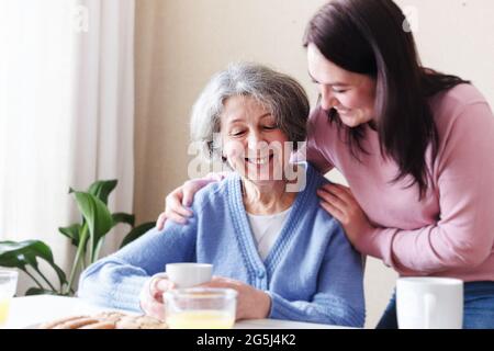 Eine Erwachsene Tochter besucht eine ältere Mutter und umarmt sie beim Frühstück sanft an den Schultern in einem gemütlichen Zuhause - das Konzept der Familienbeziehungen zwischen ihnen Stockfoto