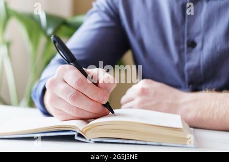 Ein Mann schreibt Notizen in einem Notizbuch mit einem Stift - EINE Männerhand macht eine Notiz in einem Notizbuch wird in Nahaufnahme gezeigt und der Hintergrund ist unscharf - das Konzept Stockfoto