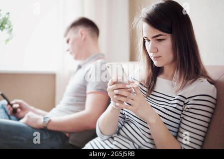 Verärgerte Ehemann und Ehefrau oder Freund und Freundin sitzen auf der Couch und schauen sich das Telefon an - verärgerte junge Menschen ignorieren sich gegenseitig und Stockfoto