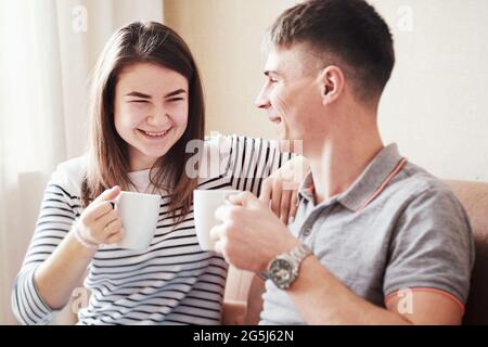 Ein Paar, das sich auf der Couch entspannt und heiße Getränke, Tee oder Kaffee trinkt - fröhliche junge Leute, die im Wohnzimmer sitzen, scherzen und sich nett unterhalten - glückliche Familie Stockfoto