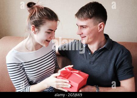 Der junge Mann umarmt und gibt seiner Frau oder Freundin eine Geschenkbox - die Frau oder Freundin nimmt an einem Jahrestag oder Feiertag ein Geschenk eines geliebten Mannes an Stockfoto