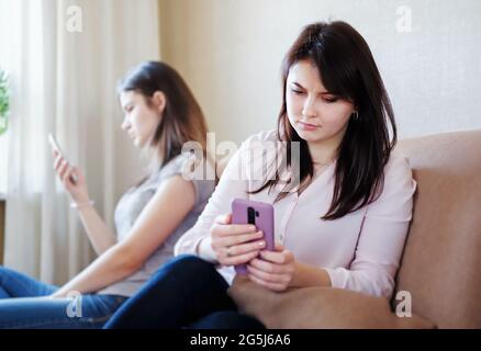 Konflikte mit Freundinnen und sie sitzen beleidigt und schauen sich ihre Telefone an - Mädchen vermissen ihre Telefone, während sie zu Hause auf der Couch sitzen - das Konzept von Stockfoto