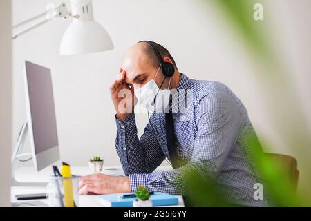 Ein Mann in einer Maske und mit einem Headset leidet unter Kopfschmerzen und Müdigkeit - Depression bei einem Callcenter-Mitarbeiter unter Bedingungen von Coronavirus - Stress Stockfoto