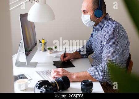 Fotograf oder Illustrator nutzt ein Grafiktablett oder retouches ein Foto – EIN digitaler Künstler, der eine Maske und Kopfhörer trägt, erstellt ein Bild auf einer Komposition Stockfoto