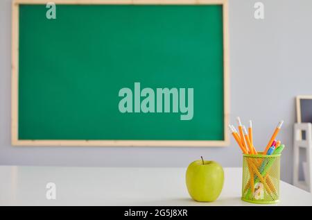 Grüner Apfel und Schreibwaren auf dem Schreibtisch des Lehrers im Klassenzimmer, leerer Raum. Obst und Schulbedarf auf dem Tisch Stockfoto