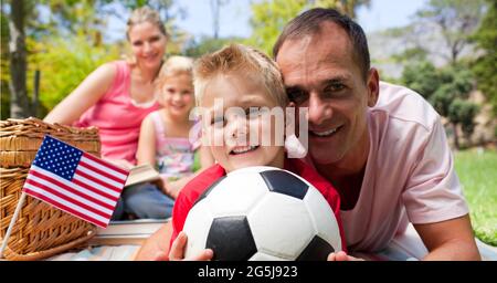 Komposition aus wehender amerikanischer Flagge, über glücklicher Familie mit Fußball beim Picknick Stockfoto