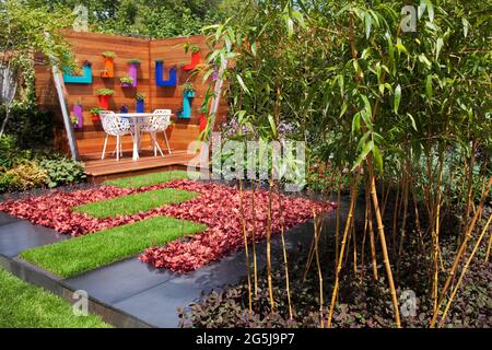 Die Pflanzen von Special Interest Nursery Garden mit einem farbenfrohen Wohnbereich im Freien mit Holzterrasse und Wand, kleinen Rasenstreifen, schwarzem Pflaster und Bambus Stockfoto