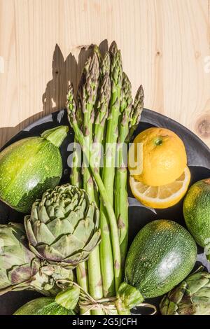 Gesundes Gemüse-Esskonzept: Ein Teller mit frischen grünen und biologischen Artischocken, Spargel, Zucchini und gelbreifen Zitronen auf einem Holztisch Stockfoto