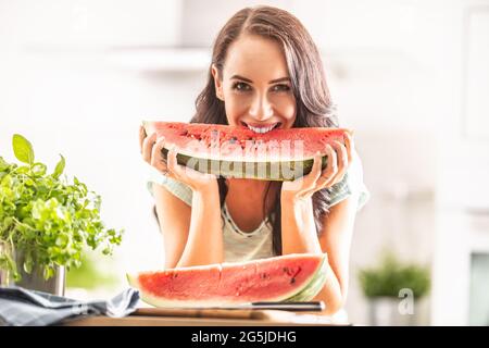 Gut aussehendes Mädchen beißt lächelnd in das Viertel einer Wassermelone in der Küche. Stockfoto