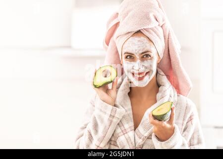 Hautpflege durch eine Frau im Kleid und Handtuch, während sie Gesichtsmaske trägt und gespaltene Avocado hält. Stockfoto