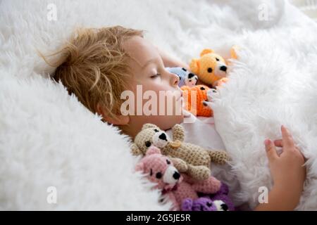 Kleines Kleinkind, niedlicher blonder Junge, der mit vielen Teddybären schläft, handgemachtes Amigurumi-Spielzeug zu Hause im Bett Stockfoto