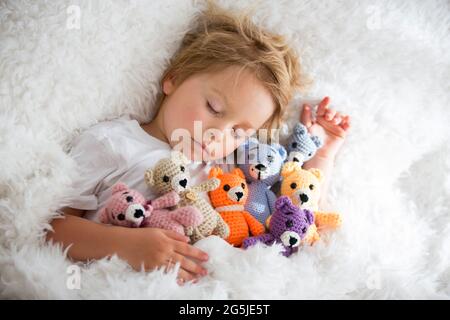 Kleines Kleinkind, niedlicher blonder Junge, der mit vielen Teddybären schläft, handgemachtes Amigurumi-Spielzeug zu Hause im Bett Stockfoto