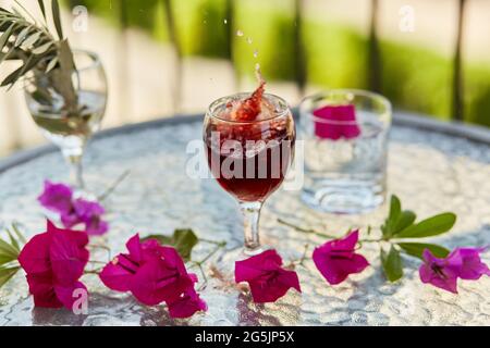 Explosion von eleganten und bunten Glas Rotwein, Wein spritzt im Glas. Sommercocktails im Hintergrund. Dekorative rosa Blüten und eine Spanke Stockfoto