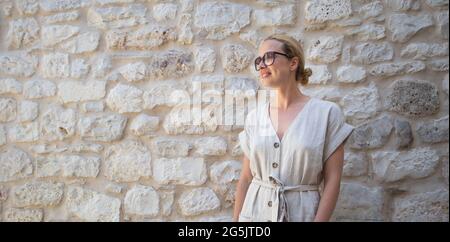 Portrait einer schönen fröhlichen blonden Frau, die ein einteiliges Sommerkleid und Sommerhut trägt und vor einer alten mittelalterlichen Steinmauer steht. Sommer Stockfoto
