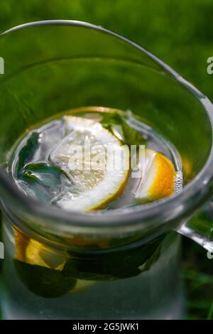 Unschärfe-Effekte aus der Nähe von Zitrone und Minze in einem Glaskrug mit Limonade auf natürlichem grünem Hintergrund. Krug mit kalten Sommercocktails oder Punsch im Freien, draußen Stockfoto