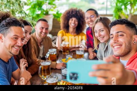 Diverse Freunde machen ein Selfie und feiern Happy Hour mit Bier und Essen in einem Bar-Restaurant. Multiethnische Menschen, die Spaß auf einer Party haben Stockfoto