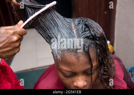Friseur Entspannen Sie das Haar auf einem afrikanischen Frauenkopf und auch mit Kamm zu strecken und gelten die Entspannungscreme durch das Haar Stockfoto