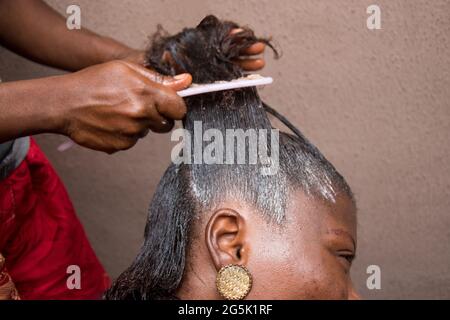 Friseur Entspannen Sie das Haar auf einem afrikanischen Frauenkopf und auch mit Kamm zu strecken und gelten die Entspannungscreme durch das Haar Stockfoto