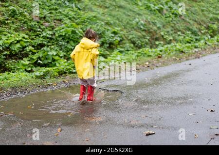 Verspielte Mädchen trägt gelben Regenmantel beim Springen in Pfütze während Regen Glückliche Kindheit Stockfoto