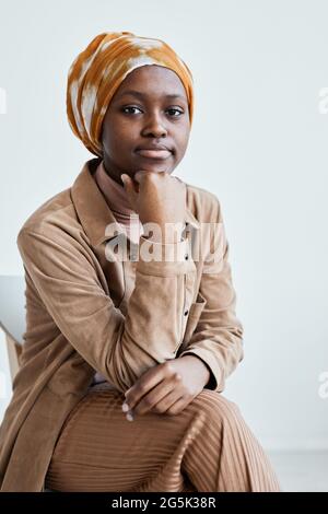 Vertikales Porträt einer modernen afroamerikanischen Frau, die die Kamera anschaut und ein Kopftuch trägt, während sie sich an einer weißen Wand posiert Stockfoto