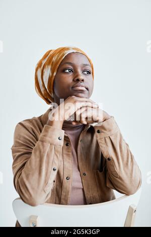 Vertikales Porträt einer modernen afroamerikanischen Frau, die wegschaut und Kopftuch trägt, während sie sich gegen die weiße Wand posiert Stockfoto