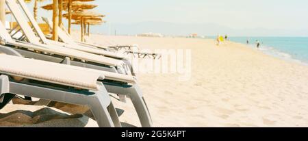 Nahaufnahme von beigefarbenen Sonnenliegen und einigen Sonnenschirmen an einem ruhigen Strand, in einem Panoramafenster, das als Webbanner oder Header verwendet werden kann Stockfoto