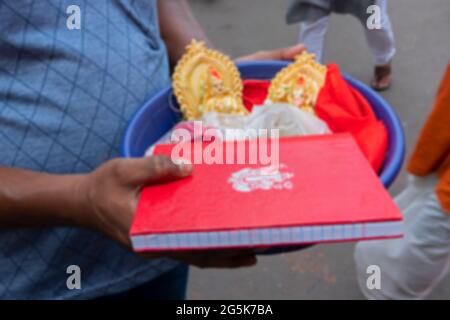 Verschwommenes Bild eines jungen Mannes, der bunte Lehmidole der Göttin Laxmi, Lord Ganesh und ein neues Notizbuch mit Ganesha-Symbol auf ihm für die Anbetung trägt Stockfoto