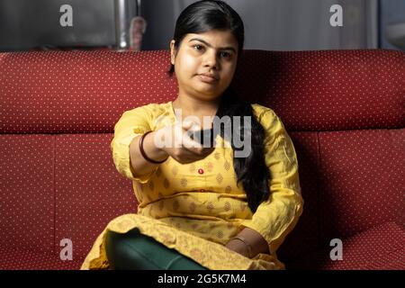 Eine indische Hausfrau, die fern in der Hand hält und auf dem Sofa sitzt Stockfoto