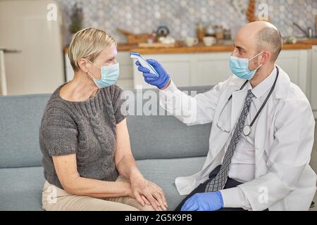 Ein Arzt misst die Temperatur einer älteren Frau in ihrem Zuhause mit einer Schutzmaske. Coronovirus-Infektionsdiagnosekonzept Stockfoto